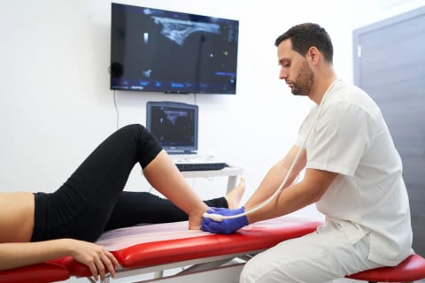 A clinician scanning the back of a patient’s ankle with an ultrasound probe, with the image shown on a screen